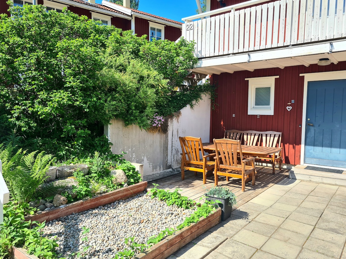 An outdoor seating area is featured with wooden chairs and a table on a deck. A pathway leads past a gravel garden with lush greenery, while the entrance to the house is marked by a blue door beneath a balcony.