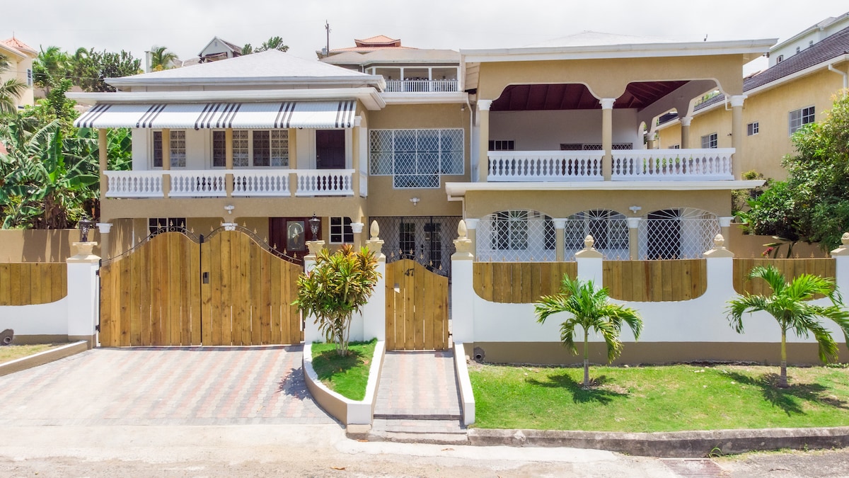 The exterior of the villa is showcased, featuring a welcoming entrance with a wooden gate and vibrant landscaping. A covered porch is visible on the upper level, adorned with decorative railings. The structure's design reflects a blend of classic and contemporary architecture.