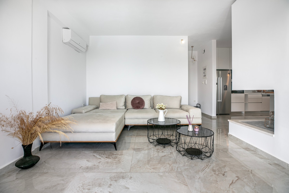 A spacious living area features a light-colored sectional sofa paired with two low, round tables. Decorative plants add a subtle touch. An open layout connects to a modern kitchen, with marble flooring reflecting natural light.