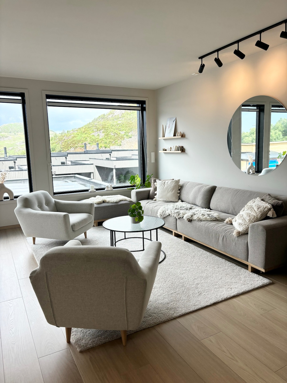 A light-filled living room features a comfortable gray sofa with soft pillows, accompanied by two matching armchairs. A round coffee table sits on a plush area rug, while a large window offers views of the surrounding mountains. Minimalist decor adds a serene touch.