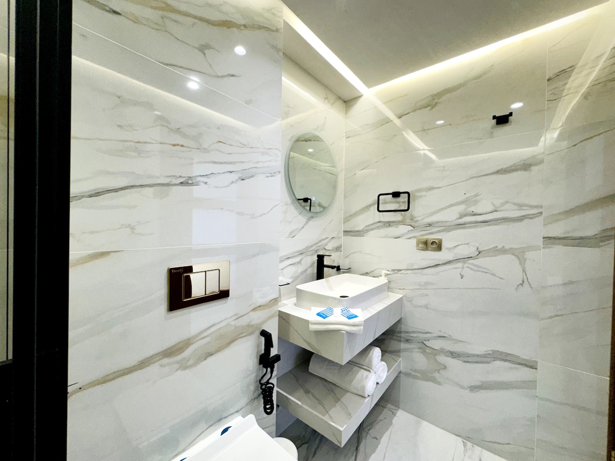 A contemporary bathroom features marble walls and a sleek countertop with a round mirror above. Towels are neatly arranged on a shelf, while modern fixtures are presented in a minimalist style, creating a clean and organized space.