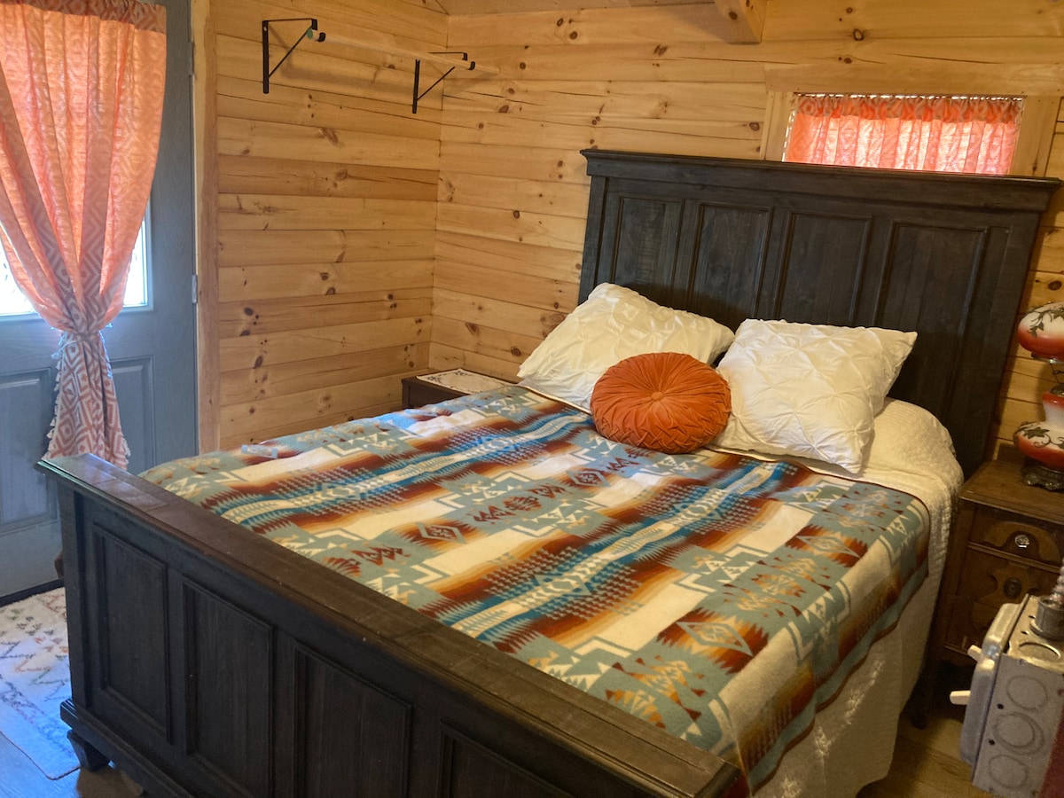 A warm and inviting sleeping area features a spacious bed adorned with a colorful patterned blanket. Two plump pillows rest at the head of the bed. Soft light filters through a partially open window, with orange curtains adding a touch of color to the wooden walls.