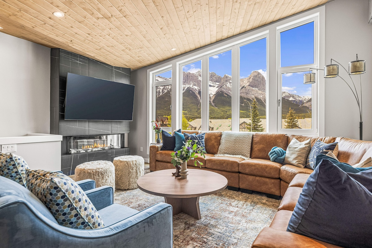 A spacious living area features a large sectional sofa and armchairs, with large windows framing mountain views. A central coffee table and a cozy fireplace invite relaxation, while soft furnishings in varied textures add comfort to the inviting atmosphere.