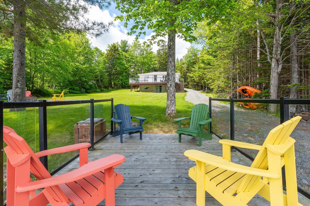 A large deck features colorful Adirondack chairs in red, blue, and yellow, providing a relaxing space. Lush green grass surrounds the cottage, framed by tall trees. The property is well-maintained, with a gravel path leading through the yard, adding to the peaceful lakeside setting.