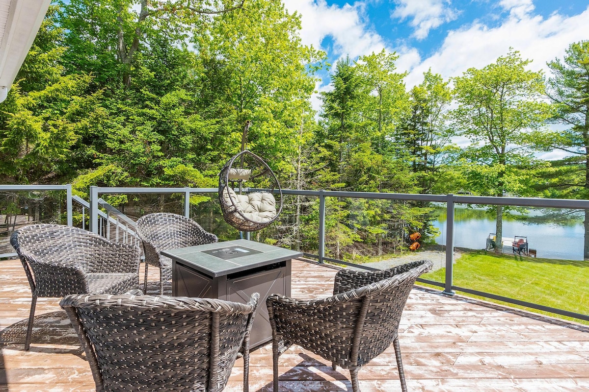 A spacious deck features a fire table surrounded by four wicker chairs, offering a comfortable seating area. Lush green trees frame the lake view in the background, enhancing the serene outdoor atmosphere.
