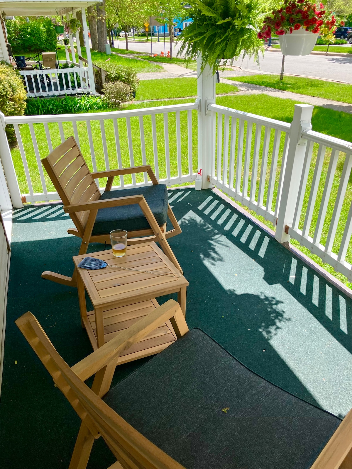 A welcoming porch space features two wooden chairs and a small side table. The green carpet and surrounding greenery create a peaceful outdoor setting, with sunlight casting shadows through the railing. Hanging planters add a touch of nature to the environment.