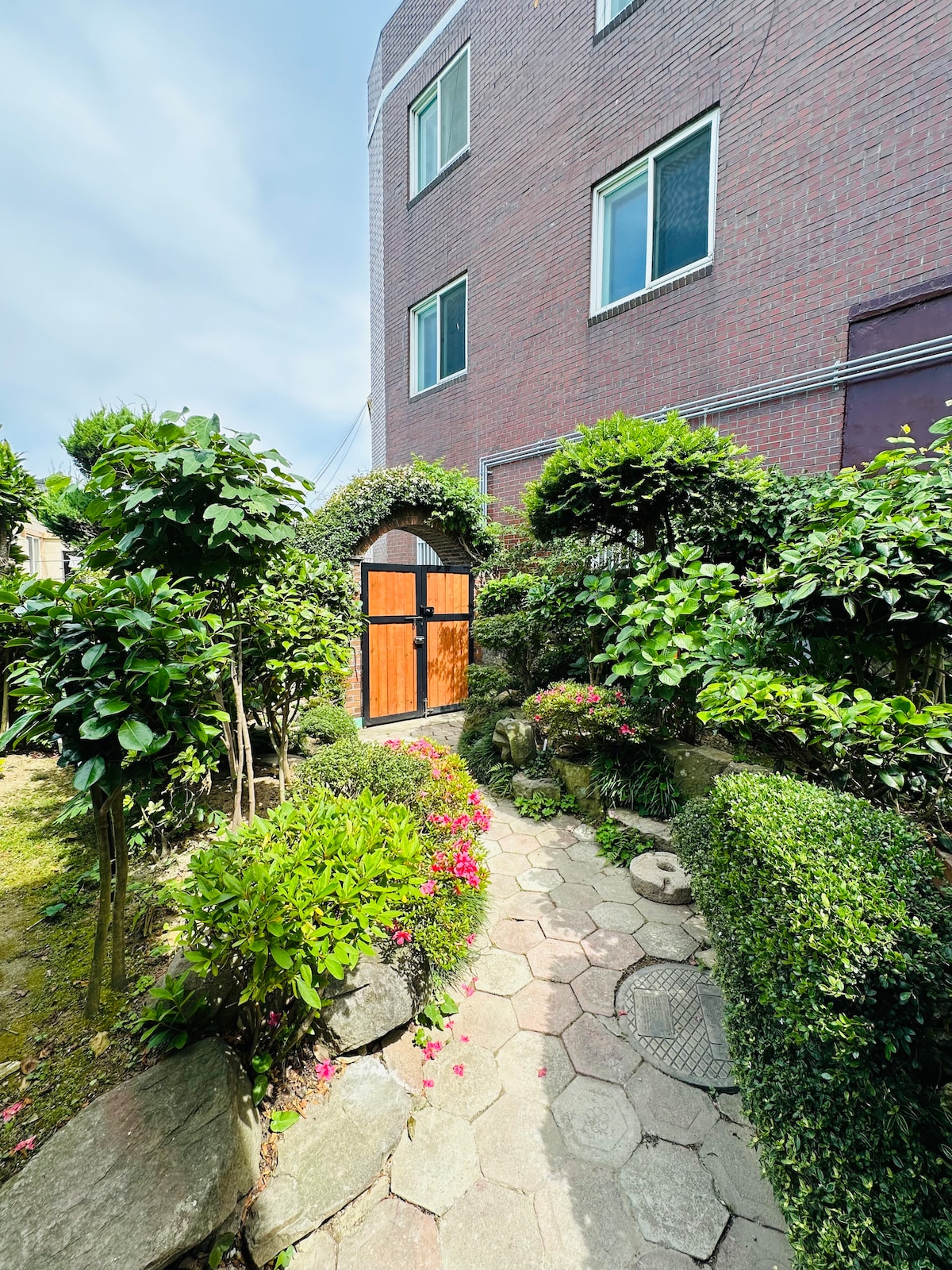 A quaint garden path leads to a wooden gate, surrounded by vibrant greenery and colorful flowers. The path is composed of hexagonal stones, creating a natural and inviting transition between the lush garden and the entryway to the property.