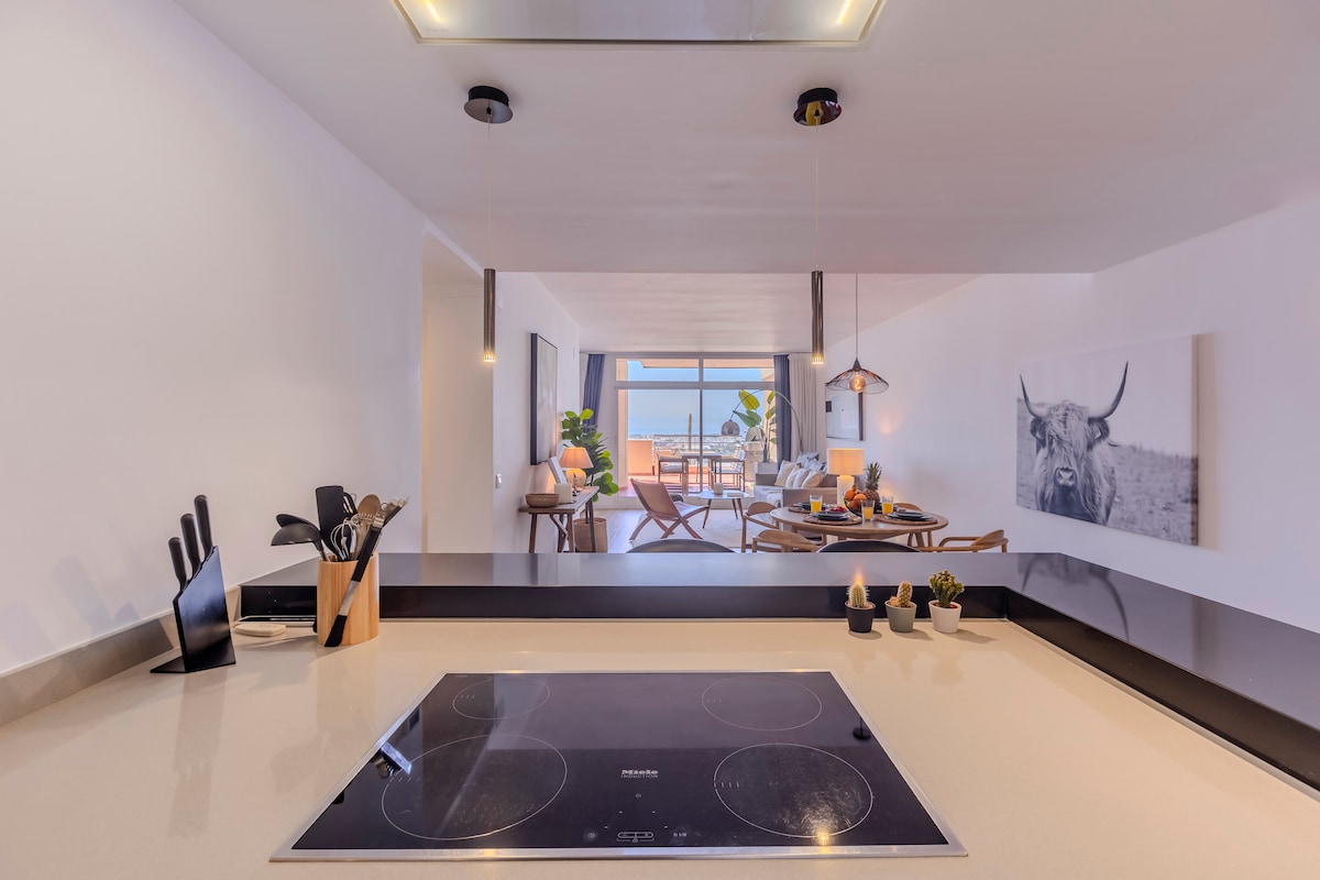 A modern kitchen is presented with a sleek countertop and an induction cooktop. Kitchen utensils are organized in a holder, while a stylish artwork of a cow adorns the wall. The space opens into a bright living area, highlighted by natural light and greenery.