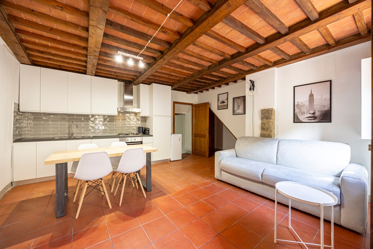 The spacious living area features a light-colored sofa accompanied by a small round table. A dining table with white chairs is positioned beside a modern kitchen with sleek cabinetry. Exposed wooden beams run across the ceiling, highlighting the rustic charm of the space.