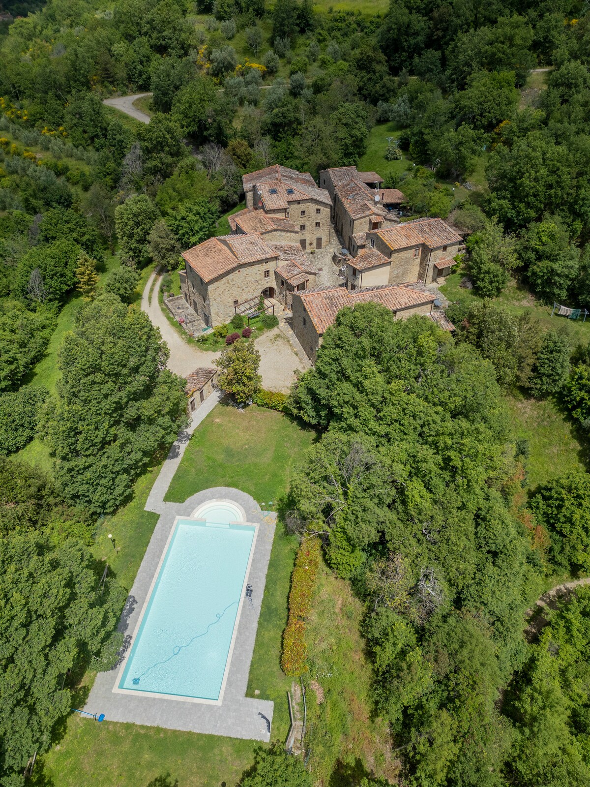 An aerial view of the historic borgo reveals a cluster of stone buildings surrounded by lush greenery. A well-maintained pool with clear water is visible in the foreground, complemented by manicured lawns and trees, creating a serene outdoor setting.