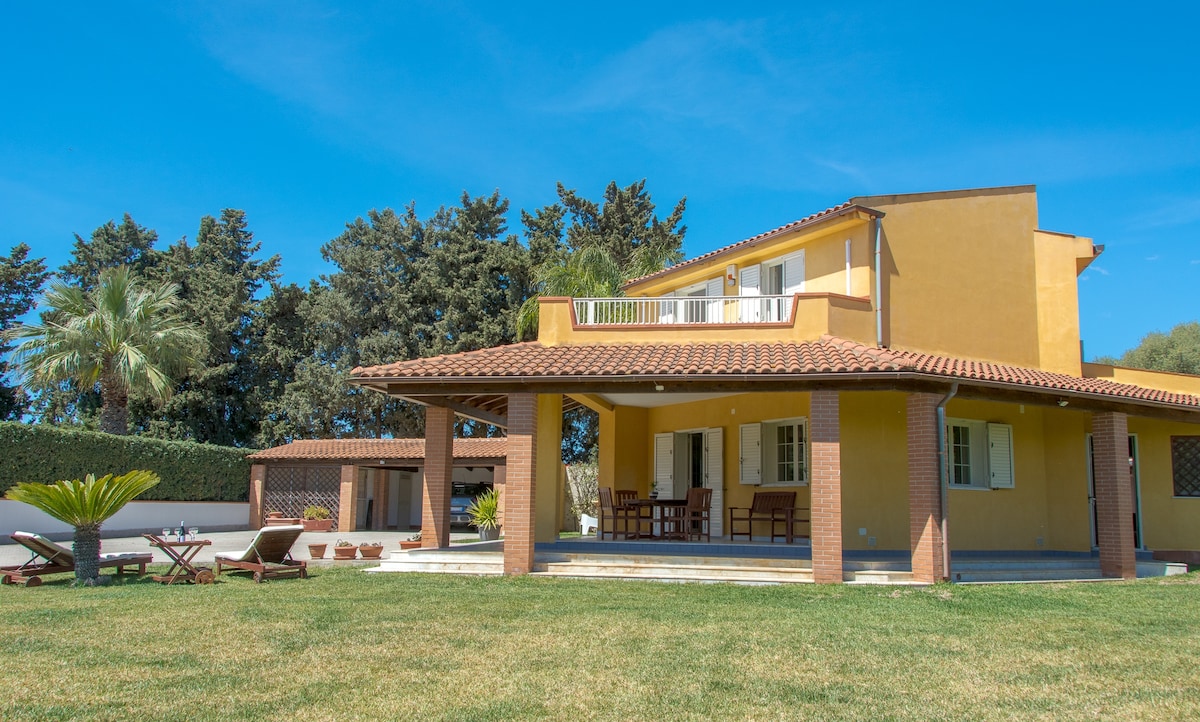 The villa is shown from a garden perspective, featuring a spacious outdoor area with a well-maintained lawn. A covered patio is highlighted, equipped with wooden furniture. The two-story structure exhibits yellow exterior walls, large windows, and a balcony overlooking the garden, surrounded by trees and greenery.