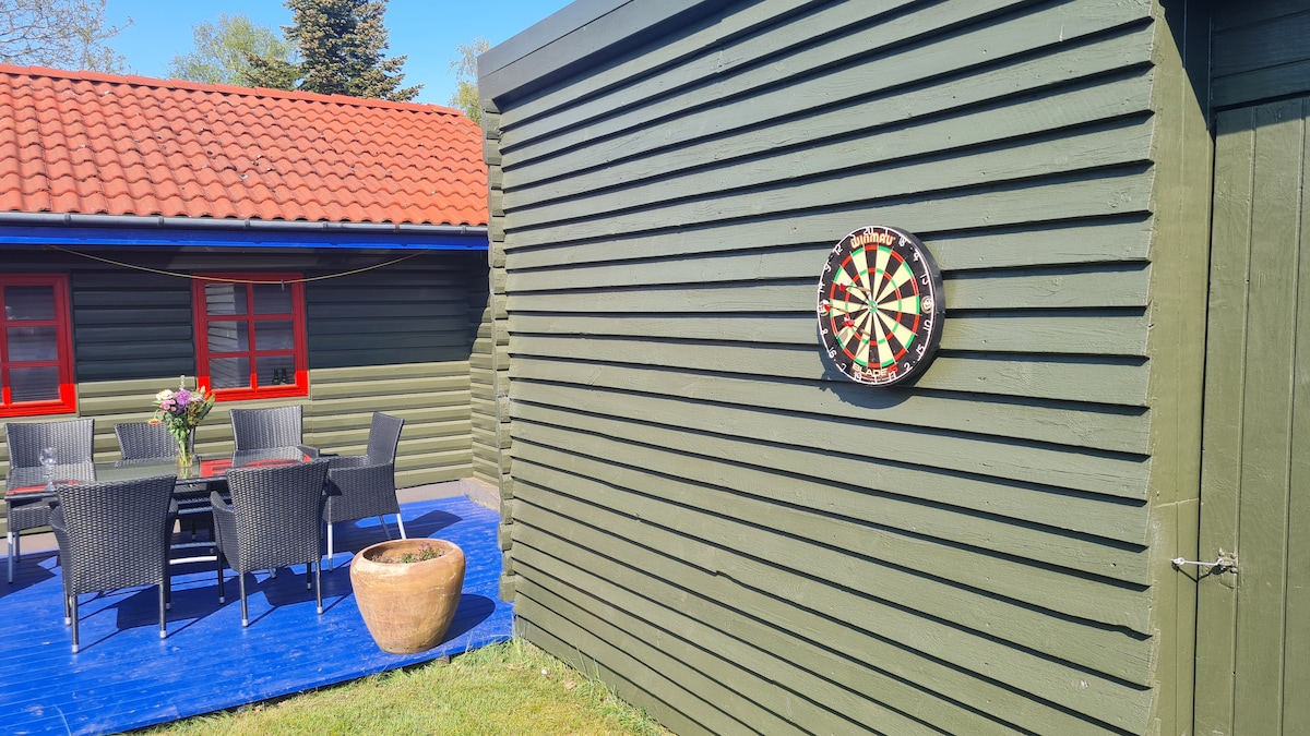 A green wooden exterior is visible, featuring a dartboard mounted on the wall. A table with black wicker chairs is nearby, accompanied by a potted plant. Red accents can be seen in the windows, enhancing the outdoor seating area.
