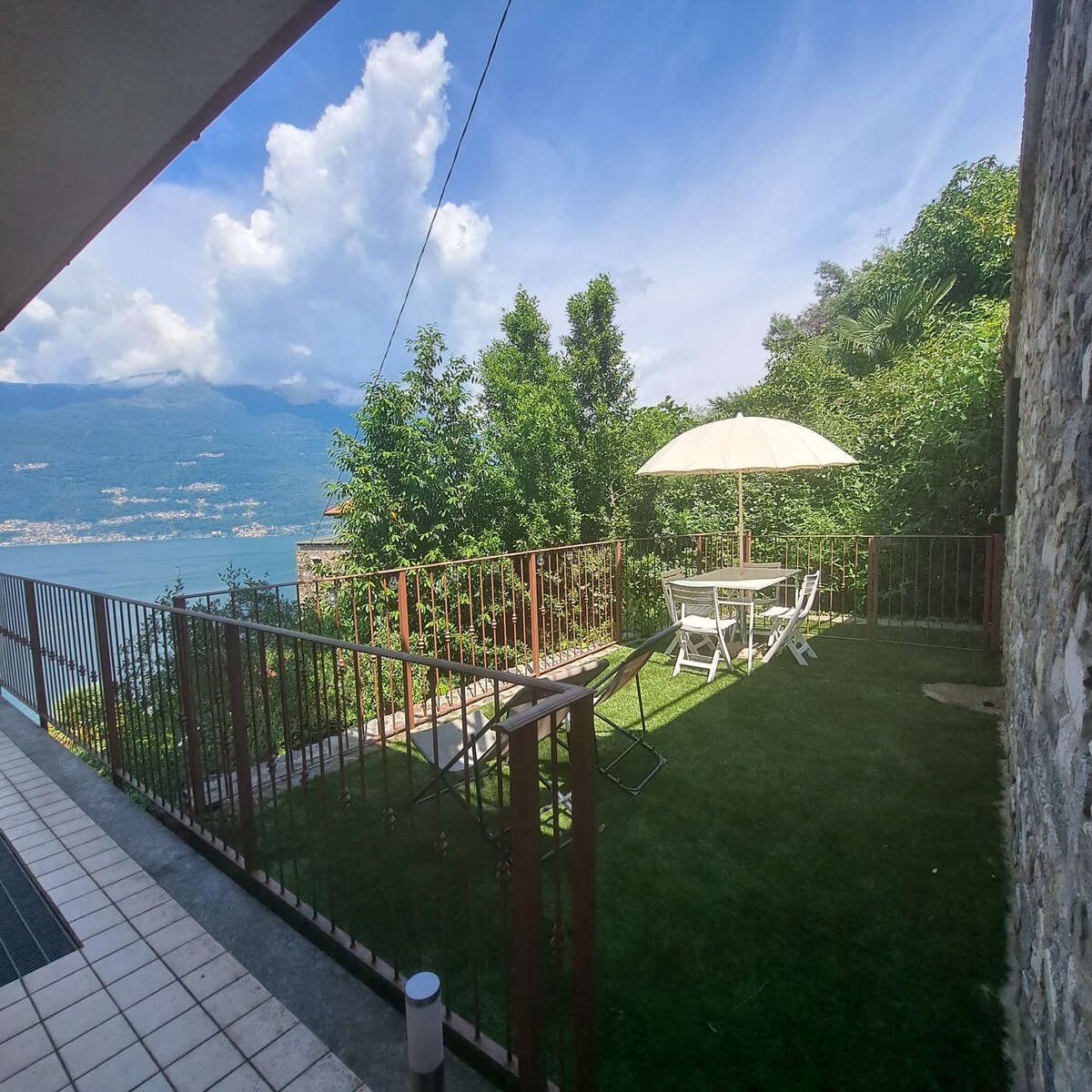 A private garden is presented with a manicured lawn, surrounded by greenery. A white patio table and chairs sit beneath a large sun umbrella, offering a relaxing space with views of the lake and mountains in the background. The area is enclosed by a low fence.