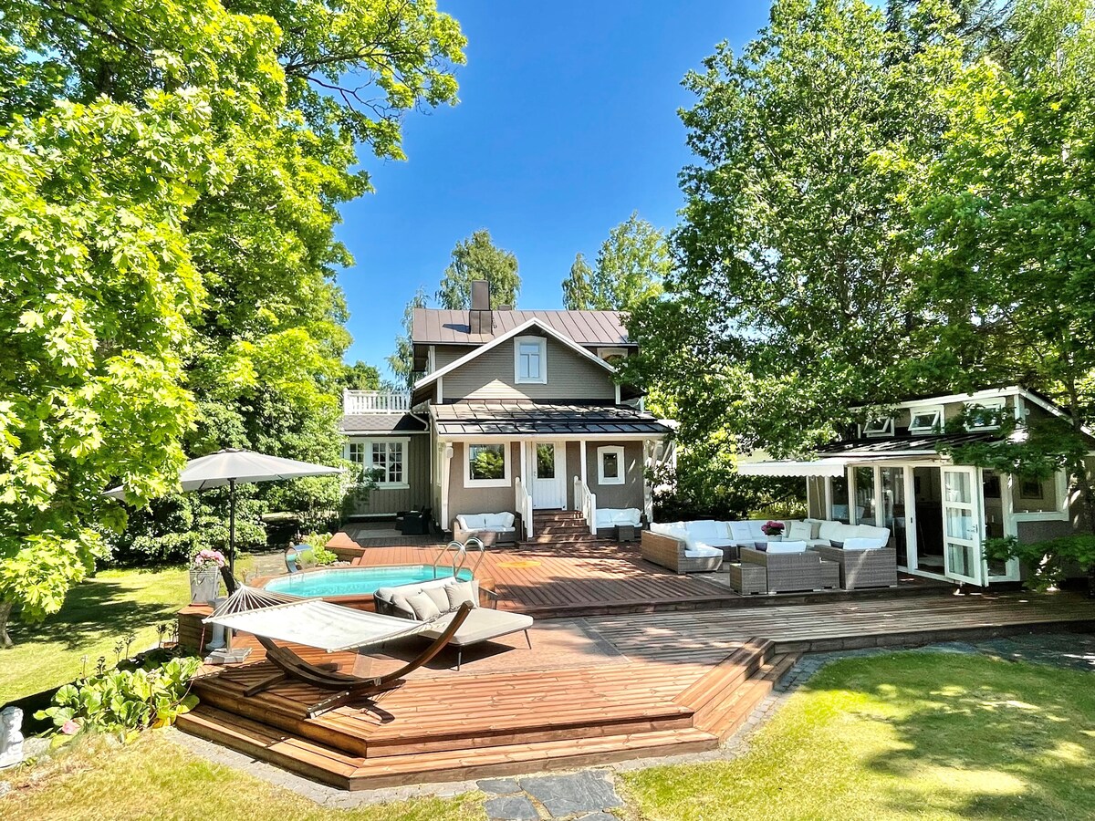 A spacious two-story house is surrounded by vibrant greenery and features a warm wooden deck. A small heated pool is positioned centrally, accompanied by lounge chairs. Outdoor seating arrangements are situated nearby, under the shade of trees and an umbrella, inviting relaxation.