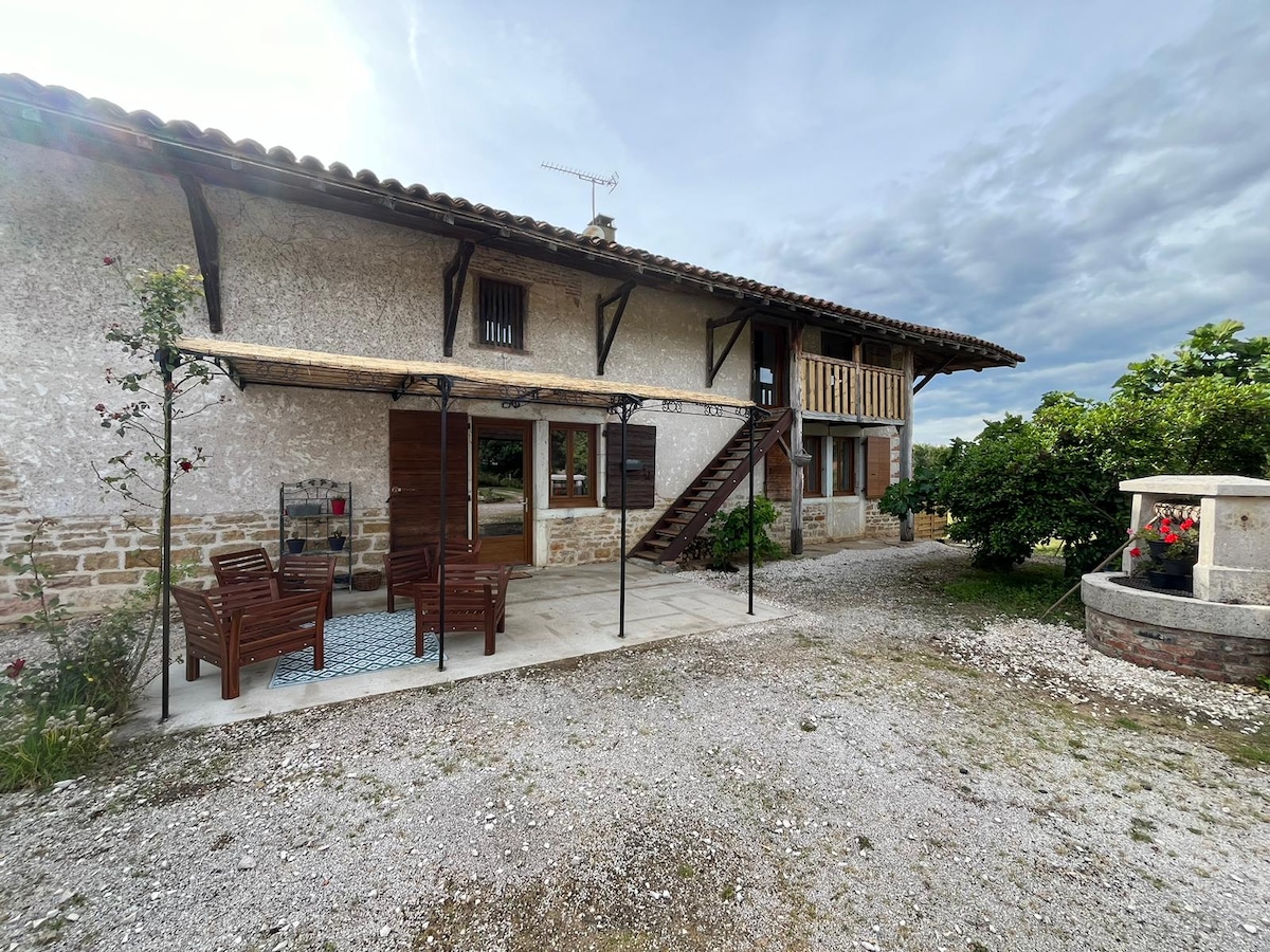 The exterior of the gîte is shown, featuring rustic stone walls and wooden elements. A small patio area with outdoor seating is present, accompanied by a staircase leading to the upper level. Lush greenery surrounds the space, creating a serene atmosphere.