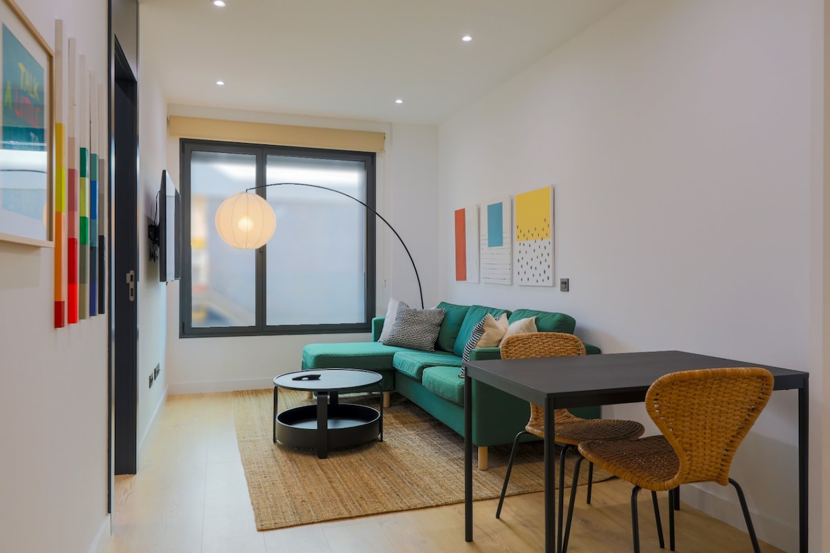 A modern living area is highlighted by a spacious green sofa, a black circular coffee table, and a round floor lamp. A neutral area rug anchors the space, while colorful wall art adds visual interest. Natural light enters through large windows adorned with simple shades.
