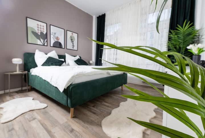 Dhomë gjumi elegante me krevat të gjelbër me susta, mbulesa me jorganë të bardhë dhe jastëkë dekorativë. Arti modern i murit dhe tavolinat pranë krevatit me abazhurë. Perdet e ndritshme dhe bimët e gjelbra krijojnë një atmosferë mikpritëse dhe komode.