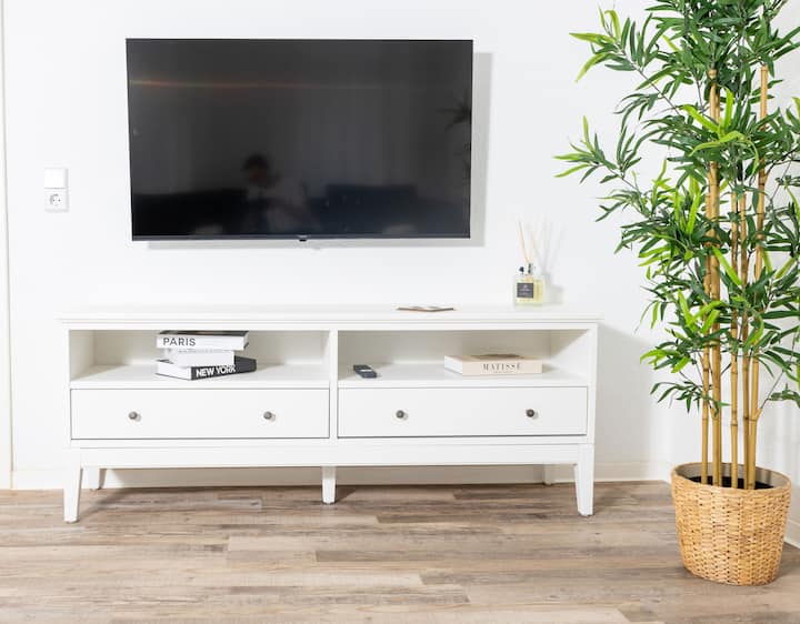 Ambient ndenjjeje i ndritshëm dhe modern me televizor me ekran të sheshtë në mur. Dërrasë e bardhë me libra dhe dekor. Bimë e madhe shtëpie e gjelbër në një shportë të endur. Dysheme e ndritshme prej druri për një atmosferë mikpritëse.