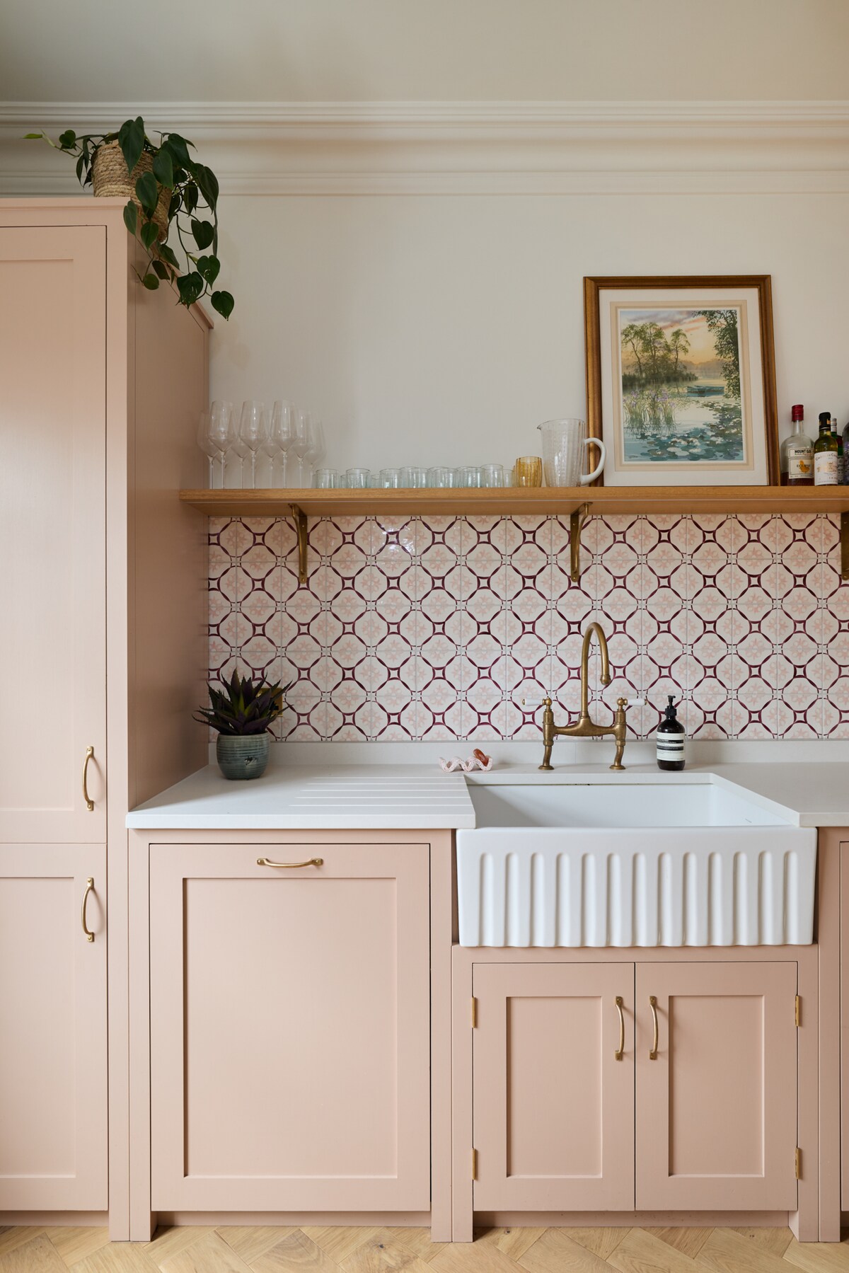 A kitchen area features soft pink cabinetry paired with a textured white farmhouse sink. A decorative backsplash with geometric patterns adds visual interest. A wooden shelf holds glassware and a plant, while a framed artwork enhances the overall design.