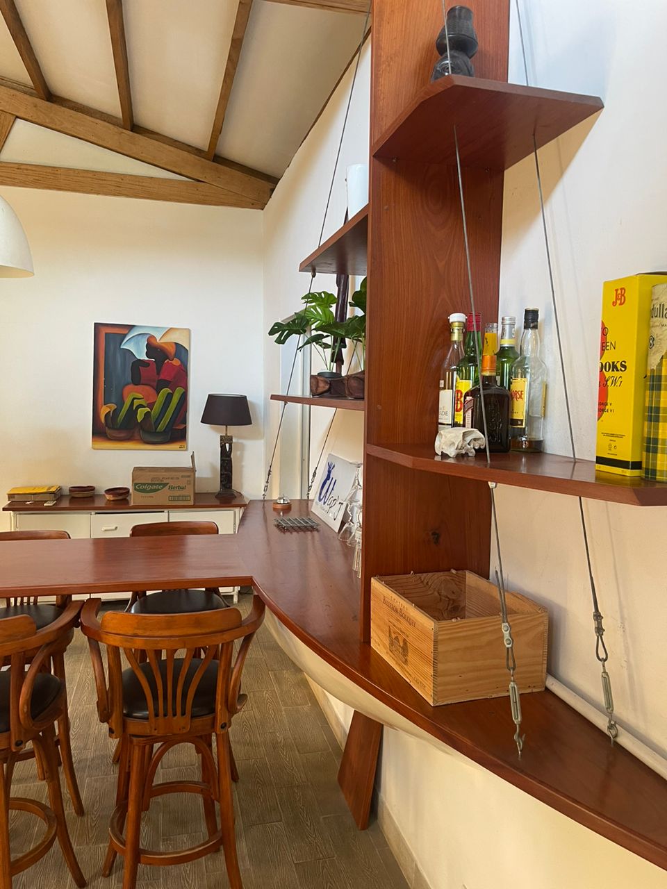 A well-appointed dining area features a wooden table surrounded by curved-back chairs. Open shelving displays a selection of bottles and decor. A vibrant piece of wall art adds a splash of color, complemented by natural light from a nearby window.