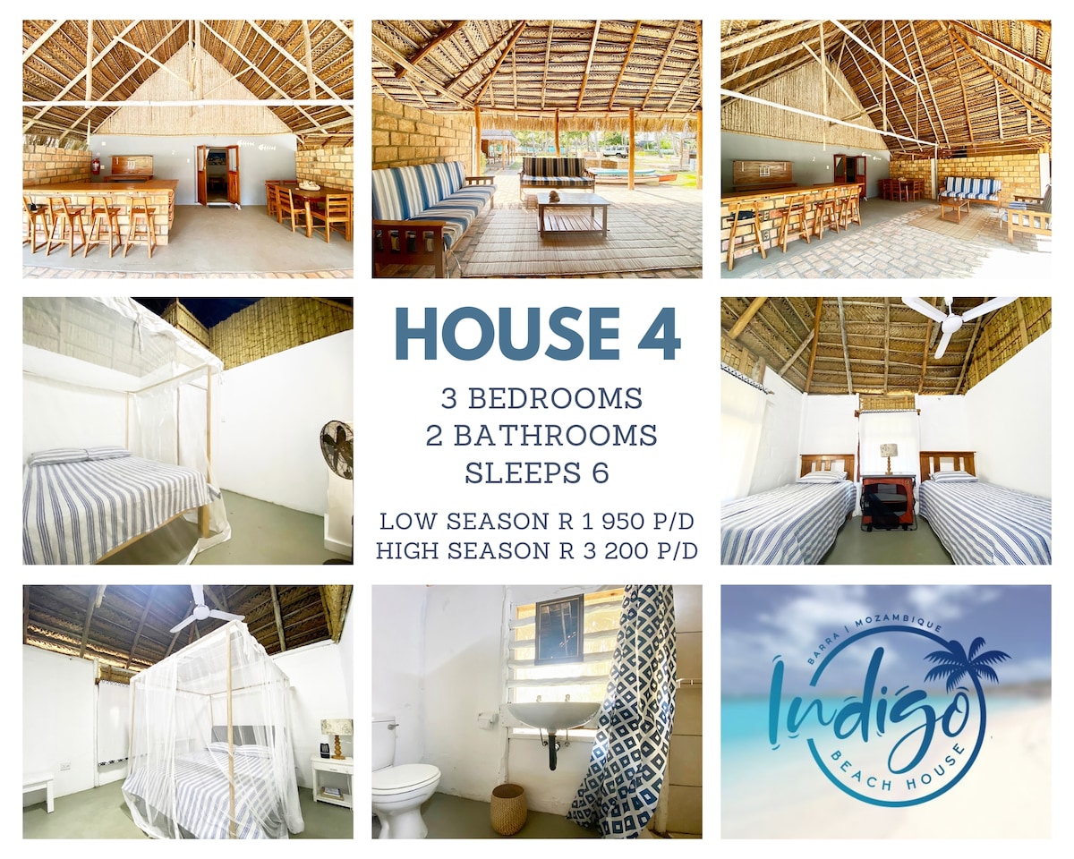 A spacious living area features wooden furniture, including a large dining table and several chairs. The room is brightened by natural light entering through large windows. Three sleeping areas are presented with comfortable beds, framed by a rustic thatched ceiling.