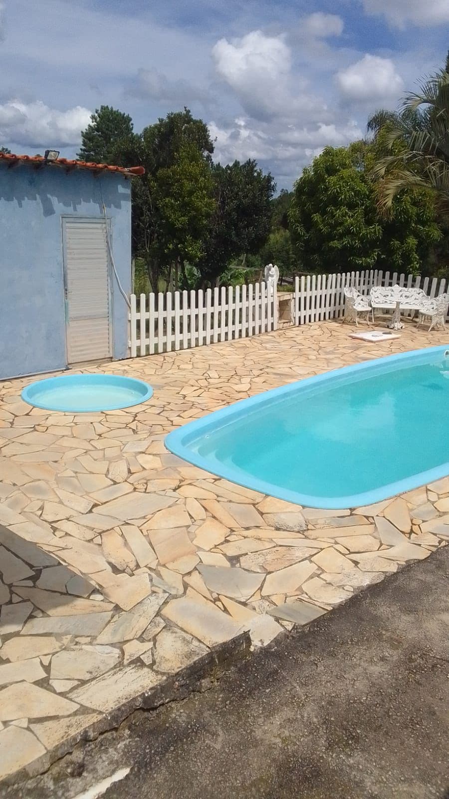 A clean swimming pool is surrounded by a stone patio, offering ample space for relaxation. A small circular area is adjacent to the pool. White fencing encircles the area, with greenery visible in the background, contributing to a peaceful outdoor setting.