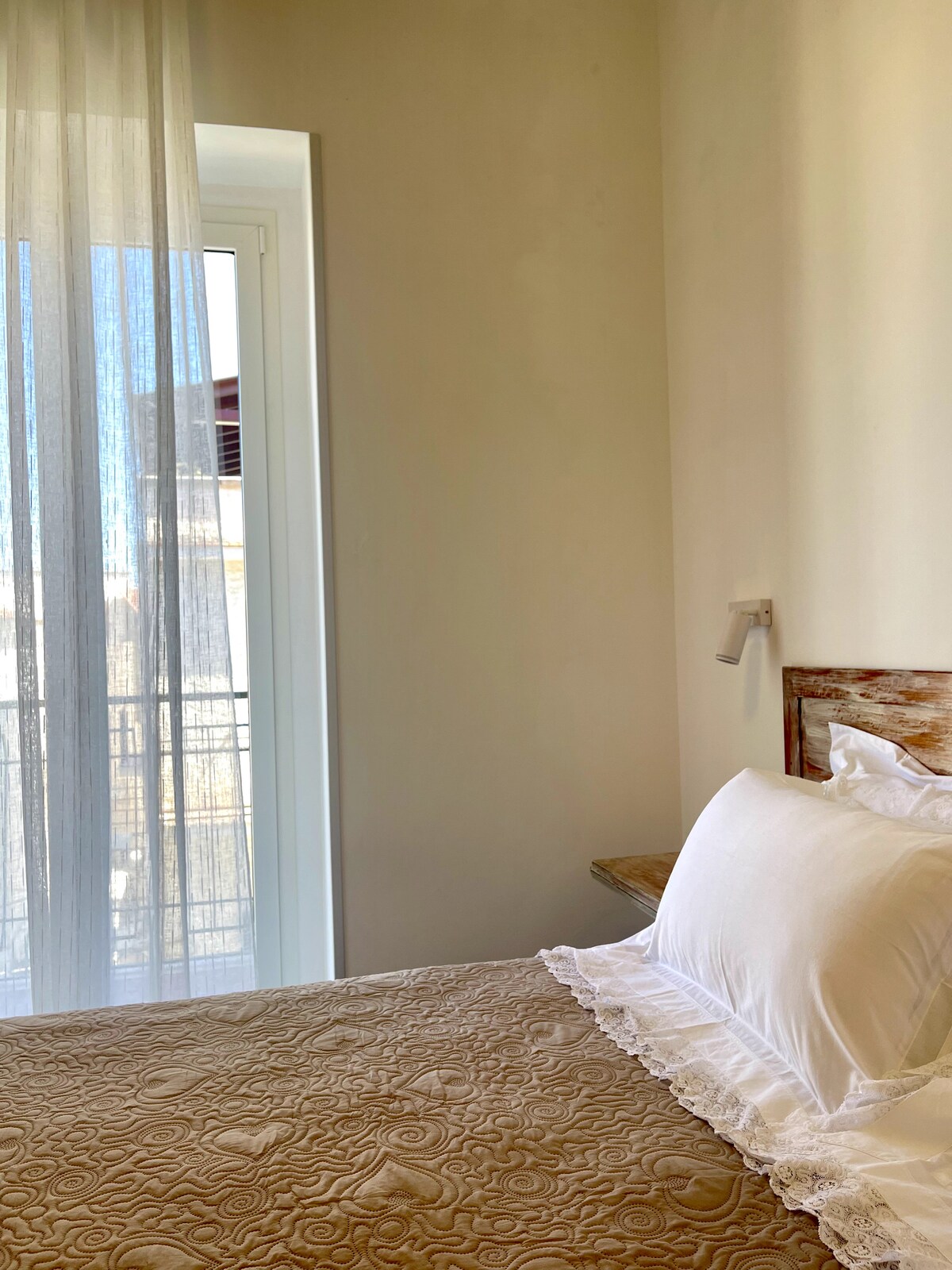 A cozy bedroom features a spacious bed with a light quilt and decorative lace trim. Soft sheer curtains filter natural light from a nearby window, leading to a small balcony with a view of the surroundings.