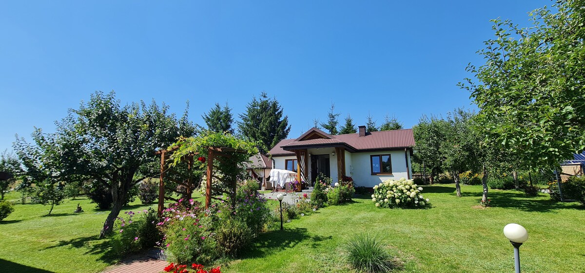 A single-story house with a brown roof is surrounded by lush greenery and vibrant flower beds. An outdoor seating area under a wooden pergola is visible, inviting relaxation. Numerous trees dot the landscape, contributing to a peaceful and spacious setting.