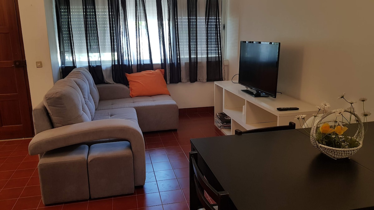 A cozy living area is furnished with a sectional sofa featuring an orange accent pillow. A television sits on a white media unit, while a dark table is positioned nearby. Large windows are dressed with sheer curtains, allowing natural light to fill the space.