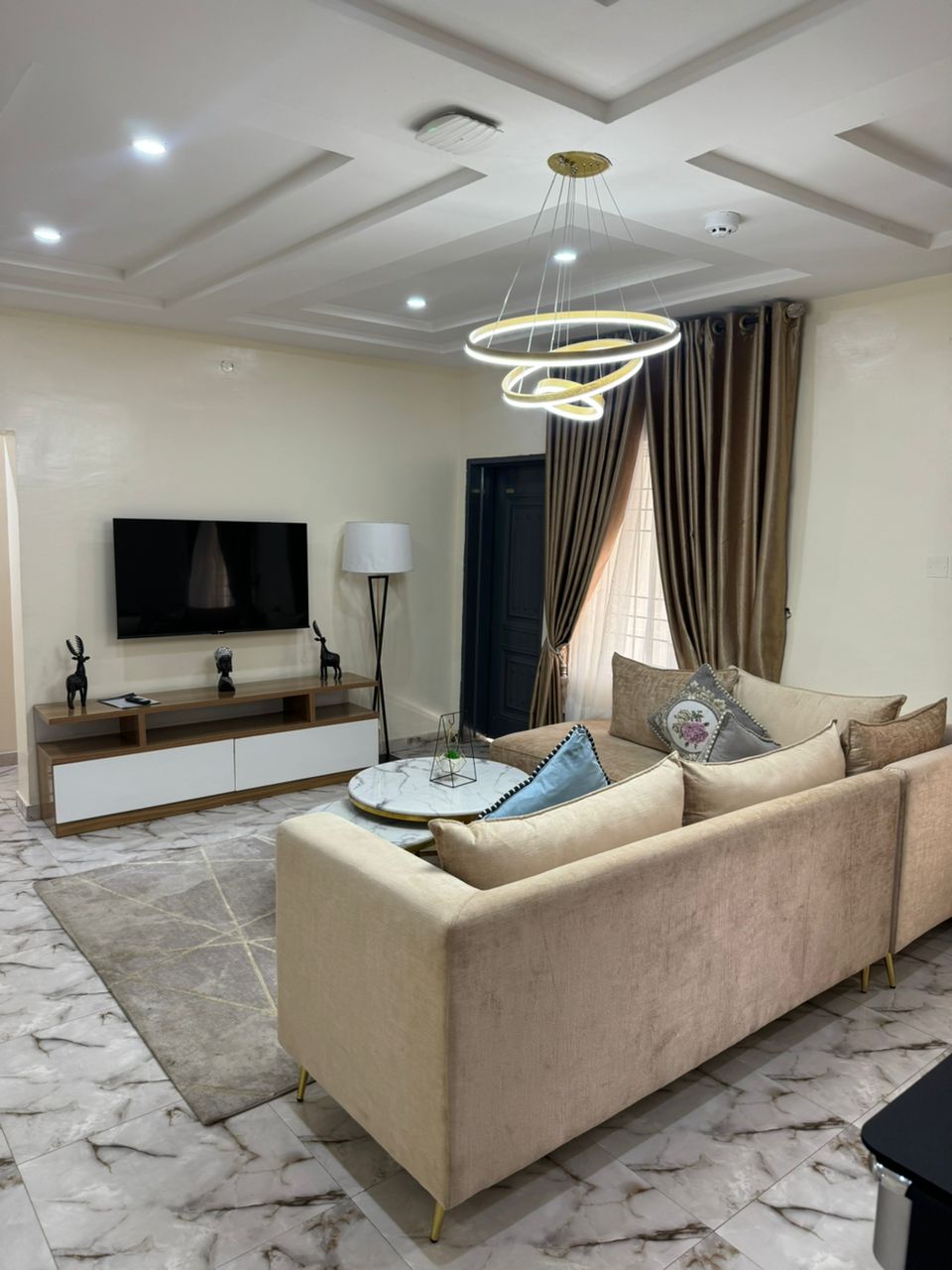 A contemporary living room showcases a beige sectional sofa adorned with decorative pillows. A round coffee table sits in front, accompanied by a flat-screen television mounted on the wall. Warm lighting is provided by a modern chandelier, and elegant curtains frame the window.
