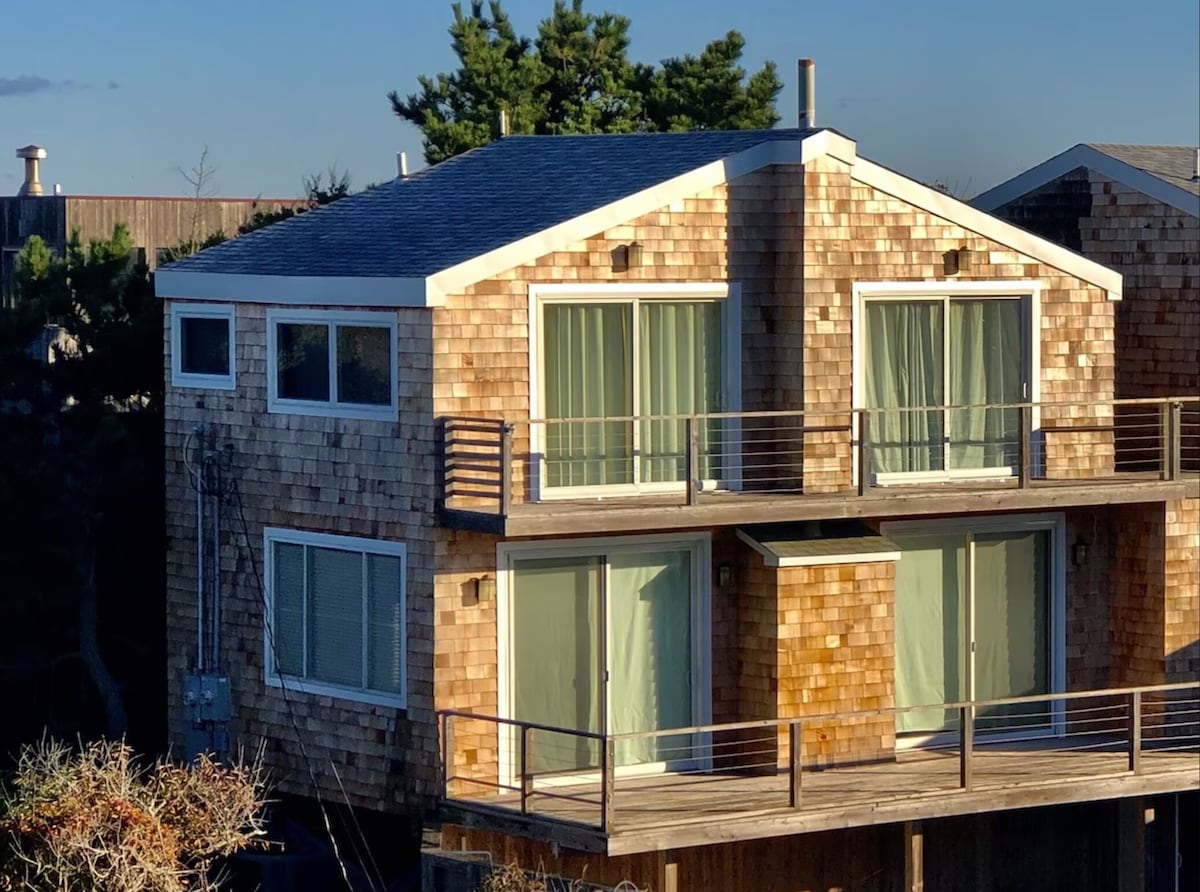 The exterior of a two-story home is shown, featuring a shingle facade and large glass sliding doors leading to balconies on both levels. Green curtains gently sway in the breeze, while greenery is visible around the property, enhancing the natural setting.