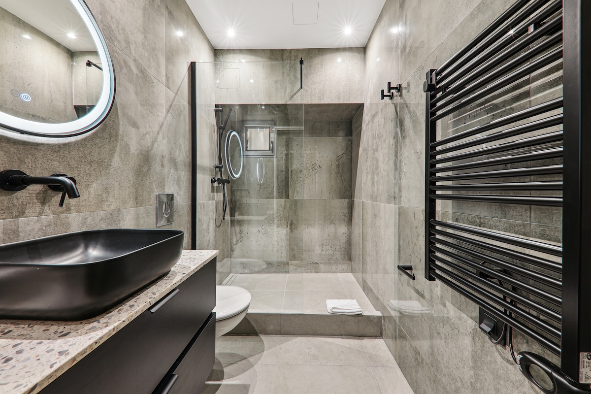 A modern bathroom features stone gray tiles and a spacious walk-in shower with a clear glass enclosure. A sleek black vanity with a round mirror is complemented by a contemporary sink. A heated towel rack is mounted on the wall, adding functionality to the design.