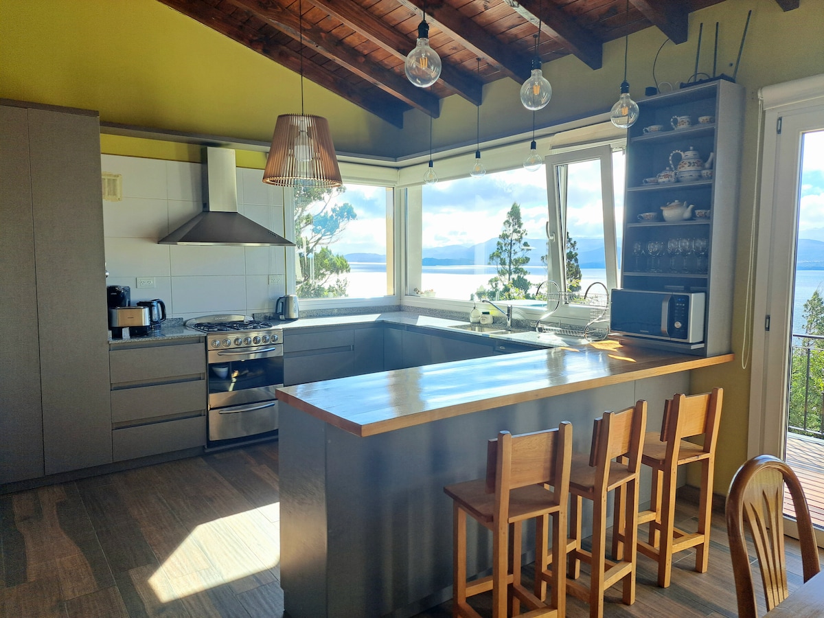 A modern kitchen is showcased with ample countertop space and sleek cabinets. A spacious island bar with wooden chairs faces large windows that offer views of the surrounding landscape. Natural light floods the area, highlighting the stainless steel appliances and open design.