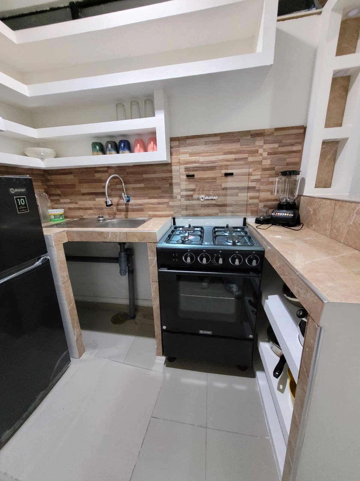 A compact kitchen area features a modern black stove with a stainless steel backsplash. Shelves display colorful dishware, while a blender and sink are conveniently positioned. The refrigerator stands to the left, enhancing the functional layout of this well-equipped cooking space.