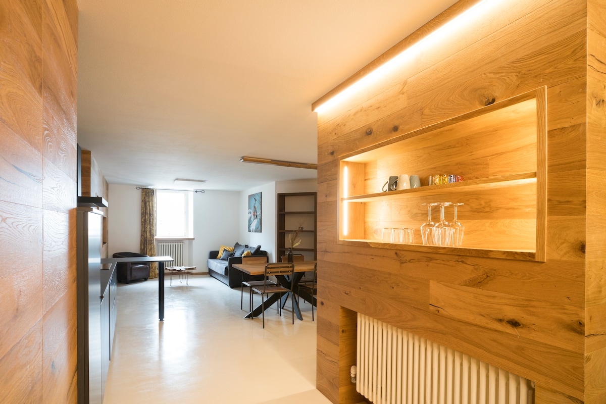 A spacious living area showcases wooden accents and modern furnishings. A wall-mounted shelf displays glassware, while a comfortable sofa and dining table are visible. Natural light filters in through a window, enhancing the clean, minimalist design of the space.