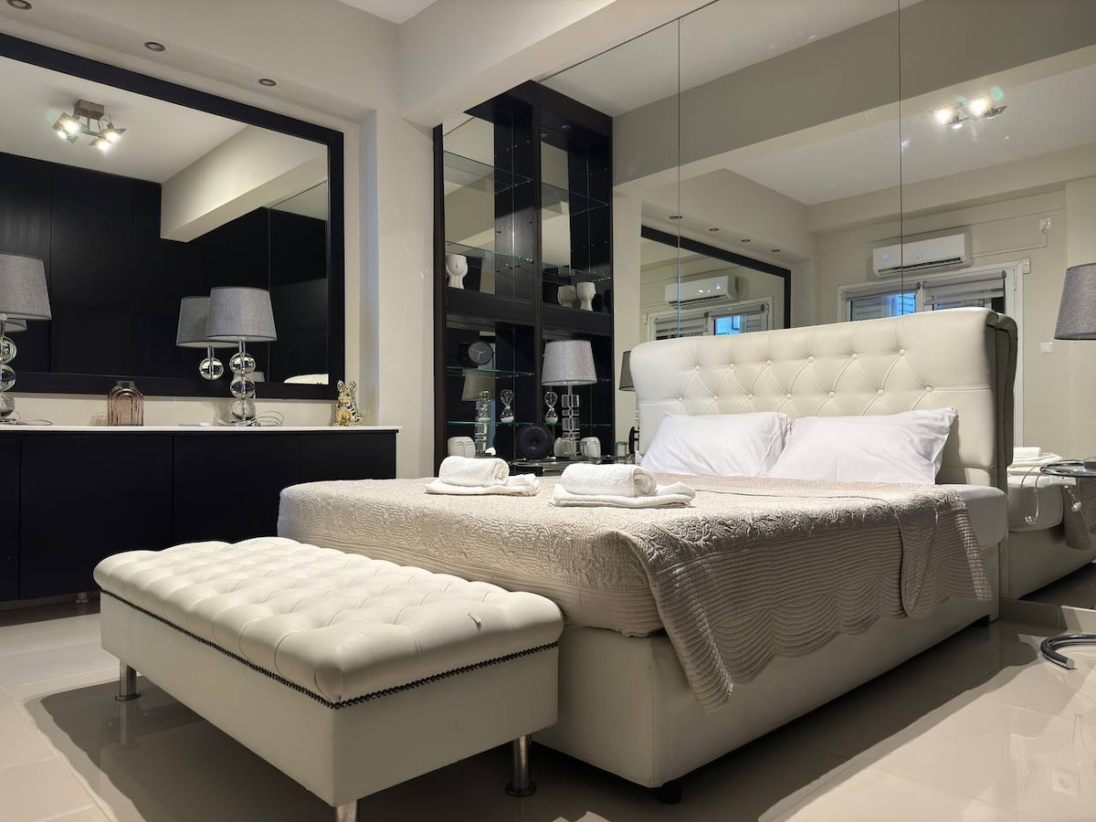 A spacious bedroom is thoughtfully arranged with a large, upholstered bed adorned with white linens. A matching ottoman sits at the foot of the bed. Modern lamps and reflective surfaces enhance the light in the room, creating a bright and stylish ambiance.