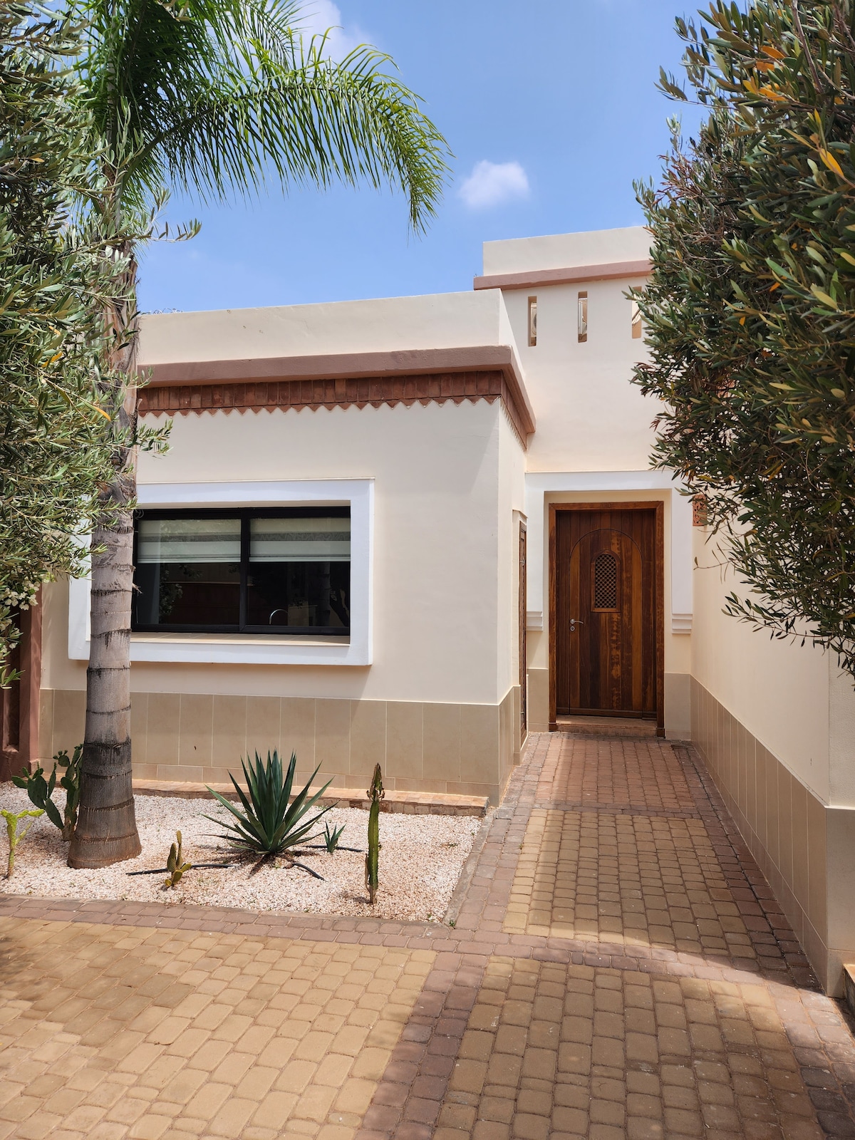 The exterior of a villa is highlighted, featuring a welcoming wooden door and a landscaped pathway. Lush greenery, including palm and cacti plants, frame the entrance. A bright window adds a touch of openness, set against a neutral color palette.