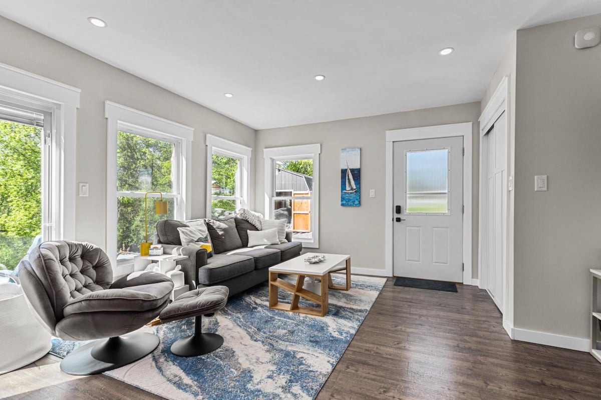 A bright living area features large windows allowing natural light to fill the space. A comfortable gray sofa is positioned alongside a unique chair, both centered around a low table on a patterned rug. The entrance door is framed by two additional windows.
