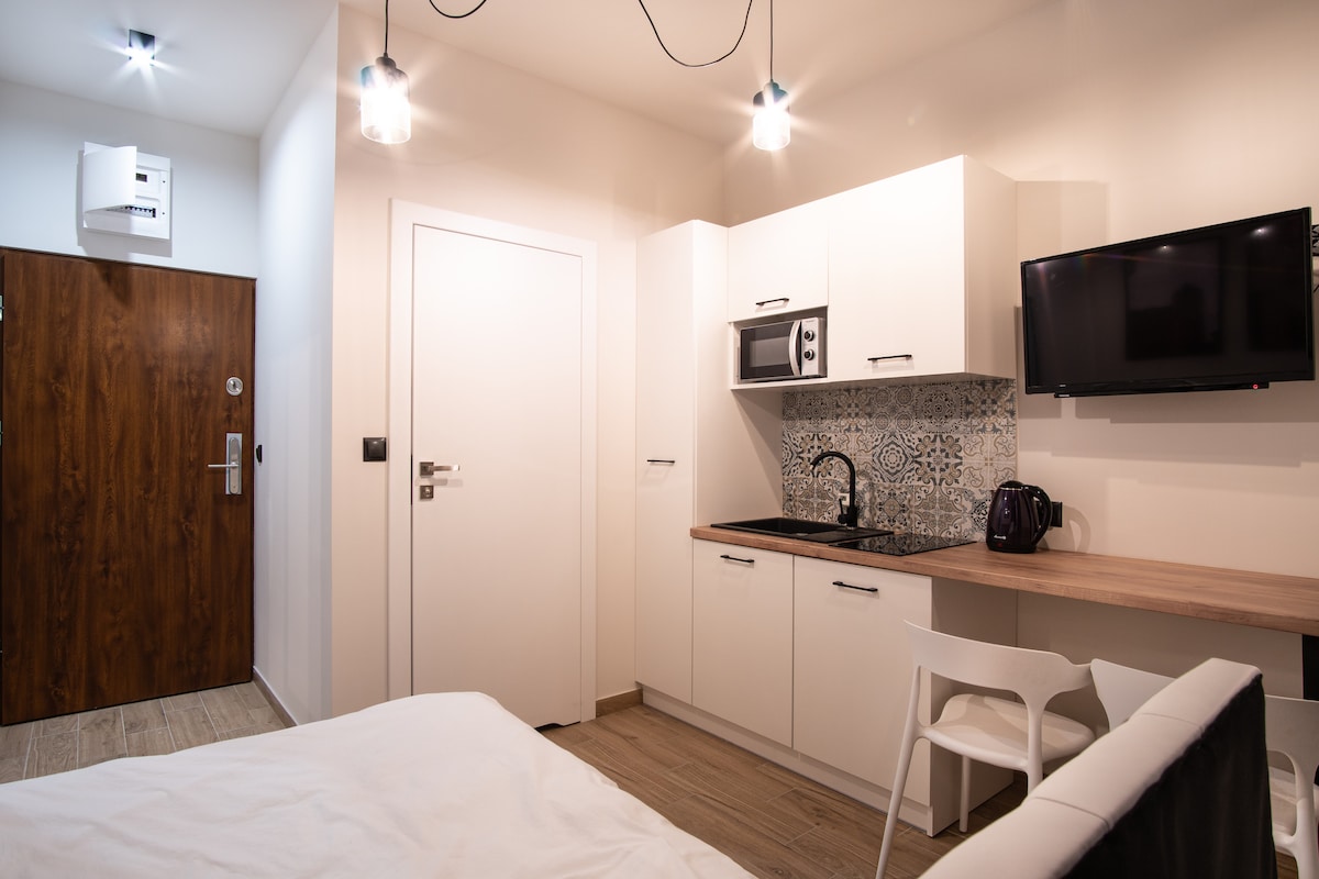 A modern studio is shown, featuring a well-equipped kitchenette with a microwave and electric kettle. A flat-screen television is mounted on the wall. The entrance door is visible, alongside a stylish wooden door. Bright lighting enhances the neutral color scheme throughout the space.