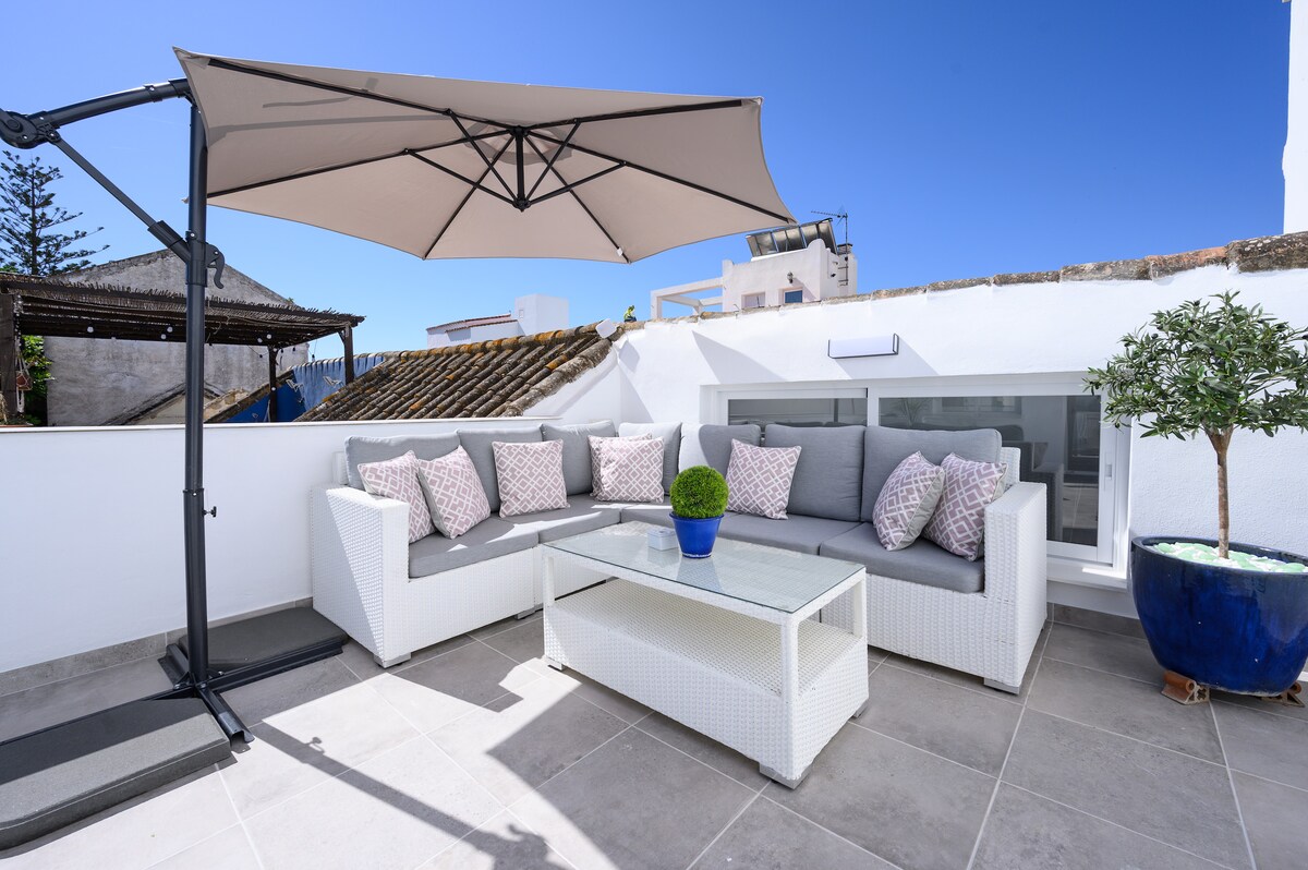 An inviting terrace area features a large white outdoor sectional sofa adorned with decorative pillows. A glass-top coffee table sits in the center, complemented by a small potted plant. An umbrella provides shade, while the backdrop includes a clear blue sky.