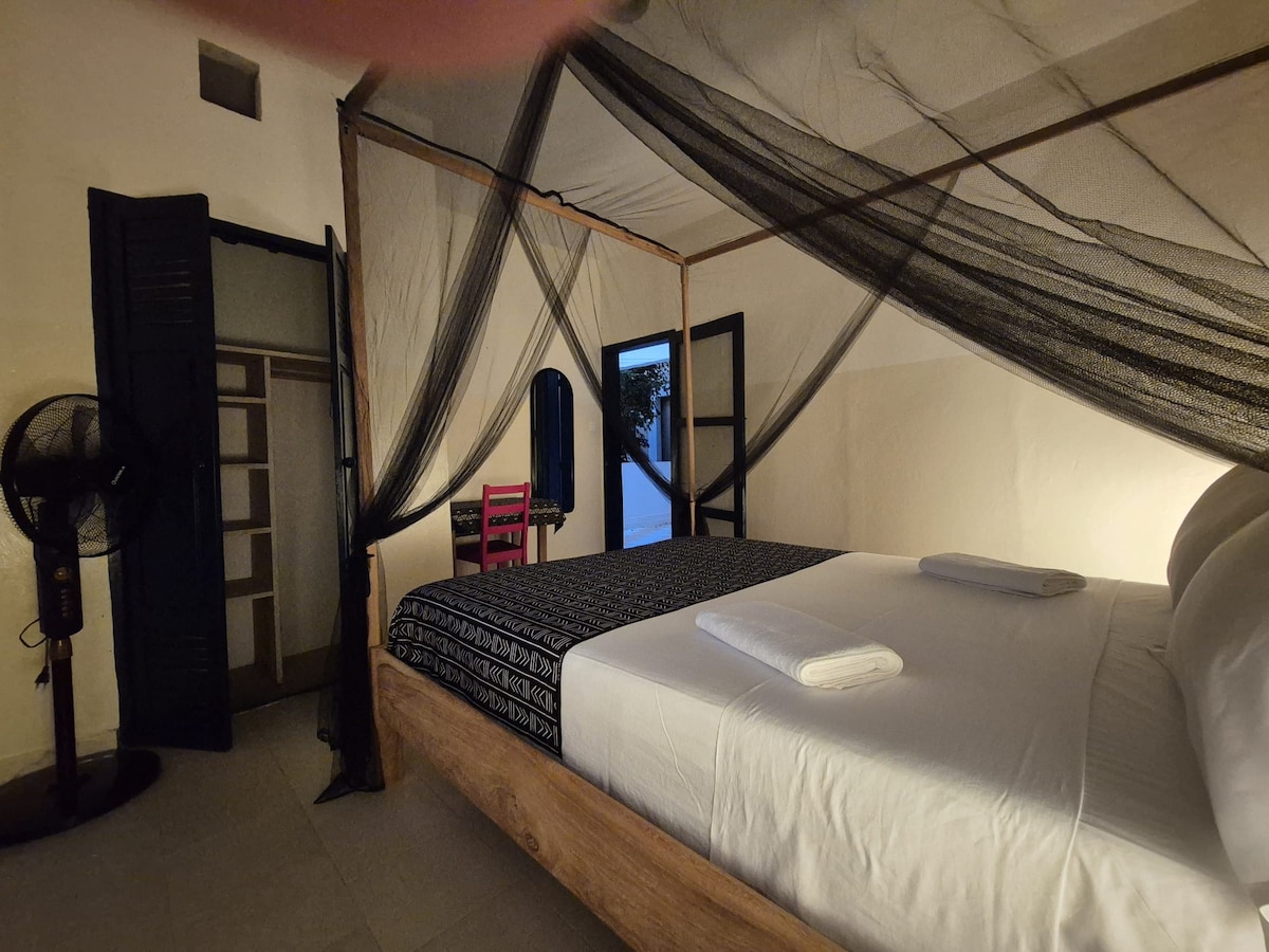 The bedroom features a spacious bed draped with a protective mosquito net. Nearby, a fan is positioned for comfort. A wooden wardrobe and two doors provide access to shared areas, with muted lighting contributing to a cozy atmosphere.