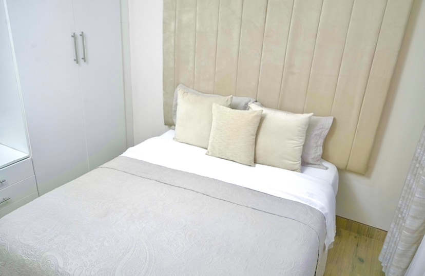 A neatly arranged sleeping area features a spacious bed dressed in soft fabrics. Four coordinating decorative pillows are positioned against a padded headboard. Light-coloured walls complement the clean design, while a wardrobe and soft drapes contribute to the serene atmosphere.