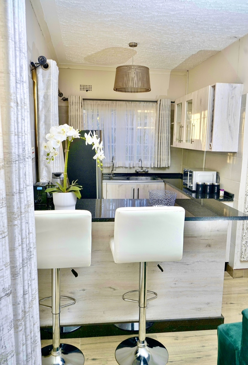 A modern kitchen space features sleek cabinetry and glossy countertops. Two white bar stools are positioned at the breakfast bar. Natural light is introduced through a window dressed with light curtains, while a potted orchid adds a touch of greenery to the design.