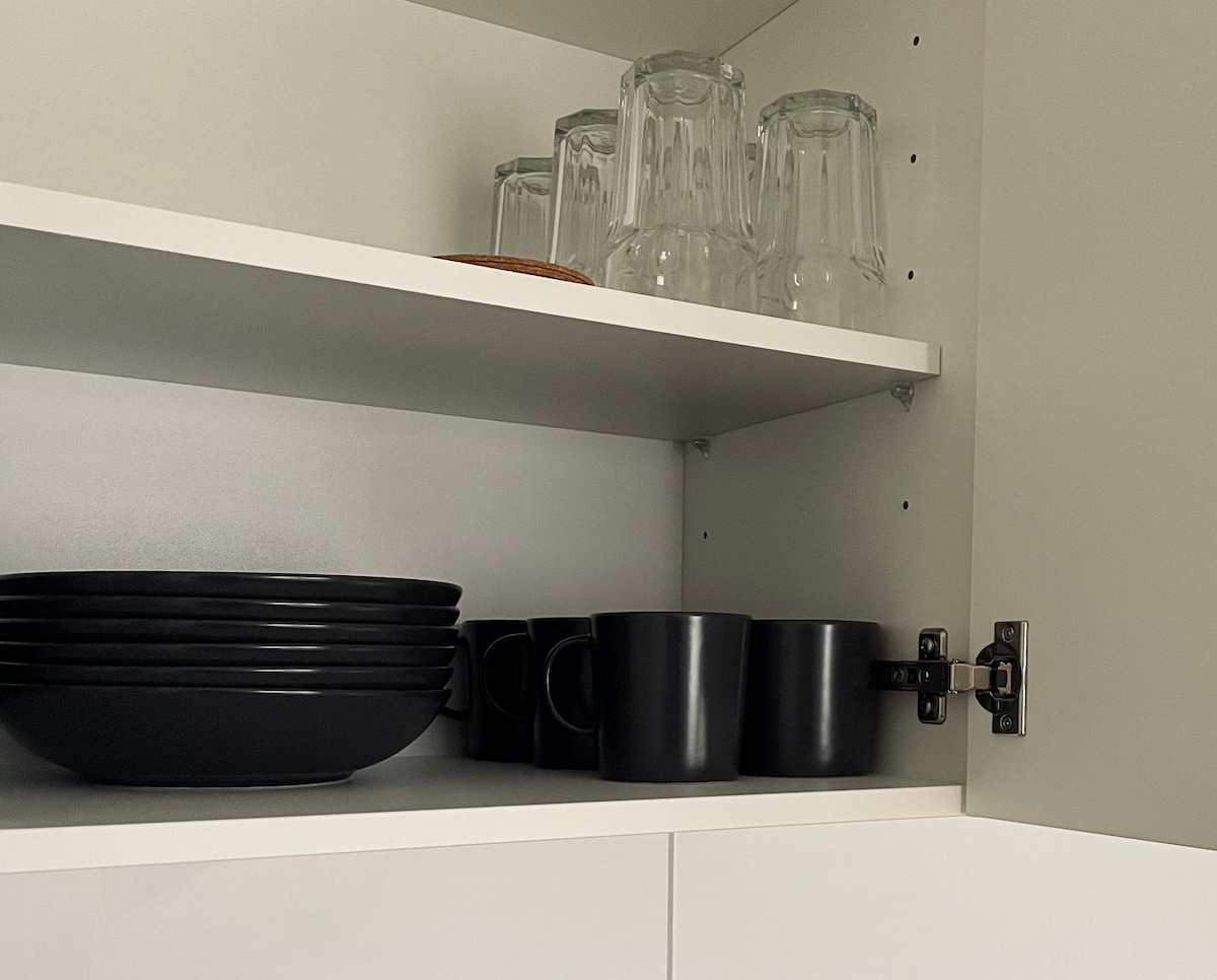 A modern kitchen cabinet is shown with an assortment of black dishware, including plates and mugs, neatly arranged. Clear glass tumblers are placed on the upper shelf, providing a functional and minimalist aesthetic.