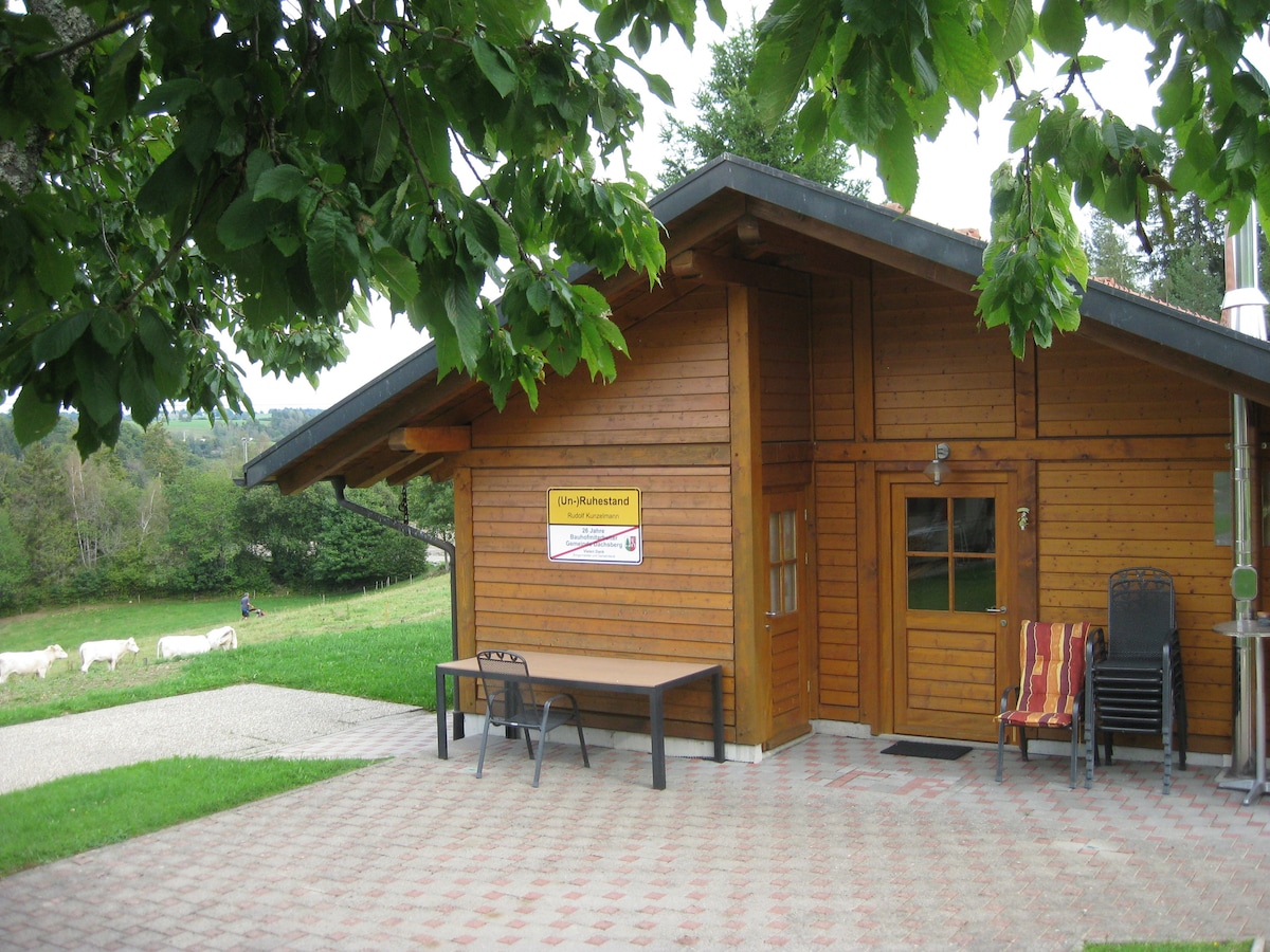 A wooden garden hut is shown, featuring a large table and chairs on a stone patio. The entrance door is framed by a gently sloping roof. In the background, a scenic view of grazing cows and green fields is visible, framed by leafy trees.