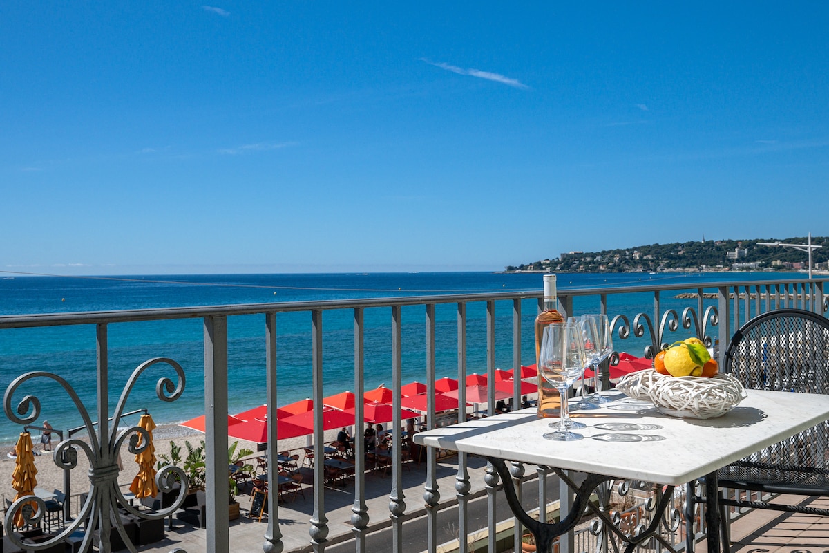 A balcony table is set with glasses and a bottle, offering a view of the sparkling blue sea and sandy beach. Red umbrellas can be seen along the shoreline, creating a vibrant contrast against the ocean. The bright sky enhances the tranquil atmosphere.