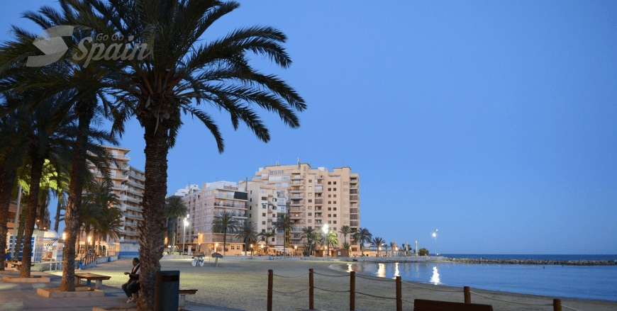 An evening view of a coastal promenade is illuminated by soft lights, with palm trees framing the scene. Low-rise buildings line the shore, reflecting the tranquil atmosphere. The calm water glistens under the twilight sky, inviting peaceful strolls along the beach.