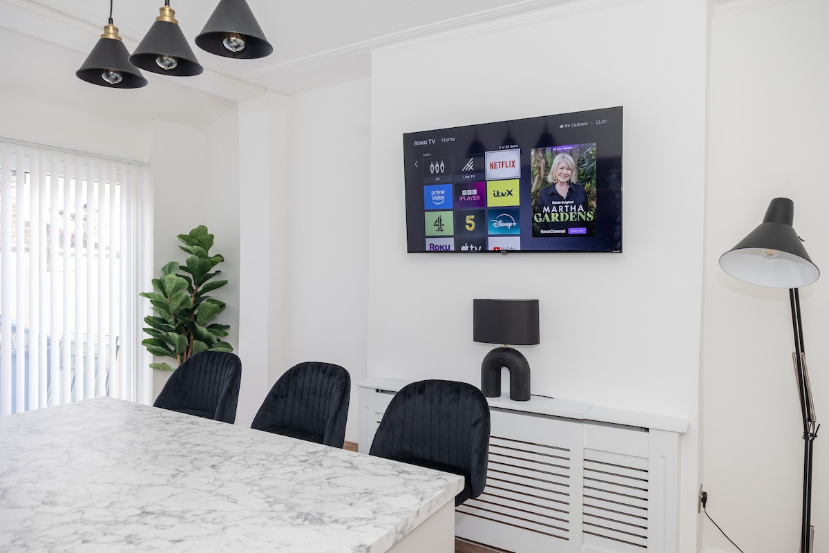 A spacious kitchen diner features a large marble dining table surrounded by four stylish black chairs. A mounted large screen Smart TV displays various streaming options, and soft natural light enters through vertical blinds. A modern lamp and a potted plant add warmth to the space.