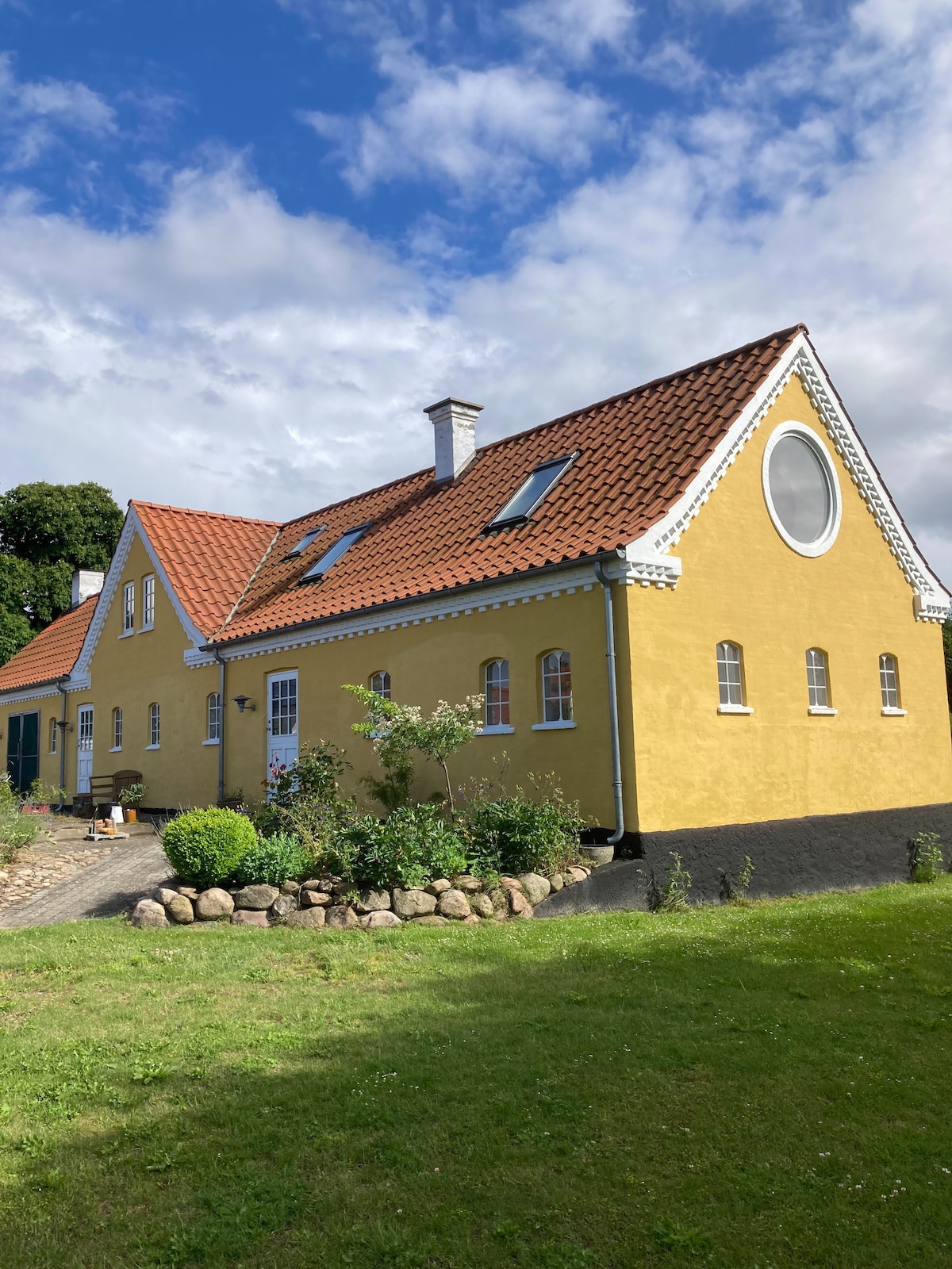 The yellow two-story house features a distinctive round window and a brick tiled roof. Well-maintained landscaping surrounds the property, including a flowerbed with stones. A grassy area is visible in the foreground, under partly cloudy skies.