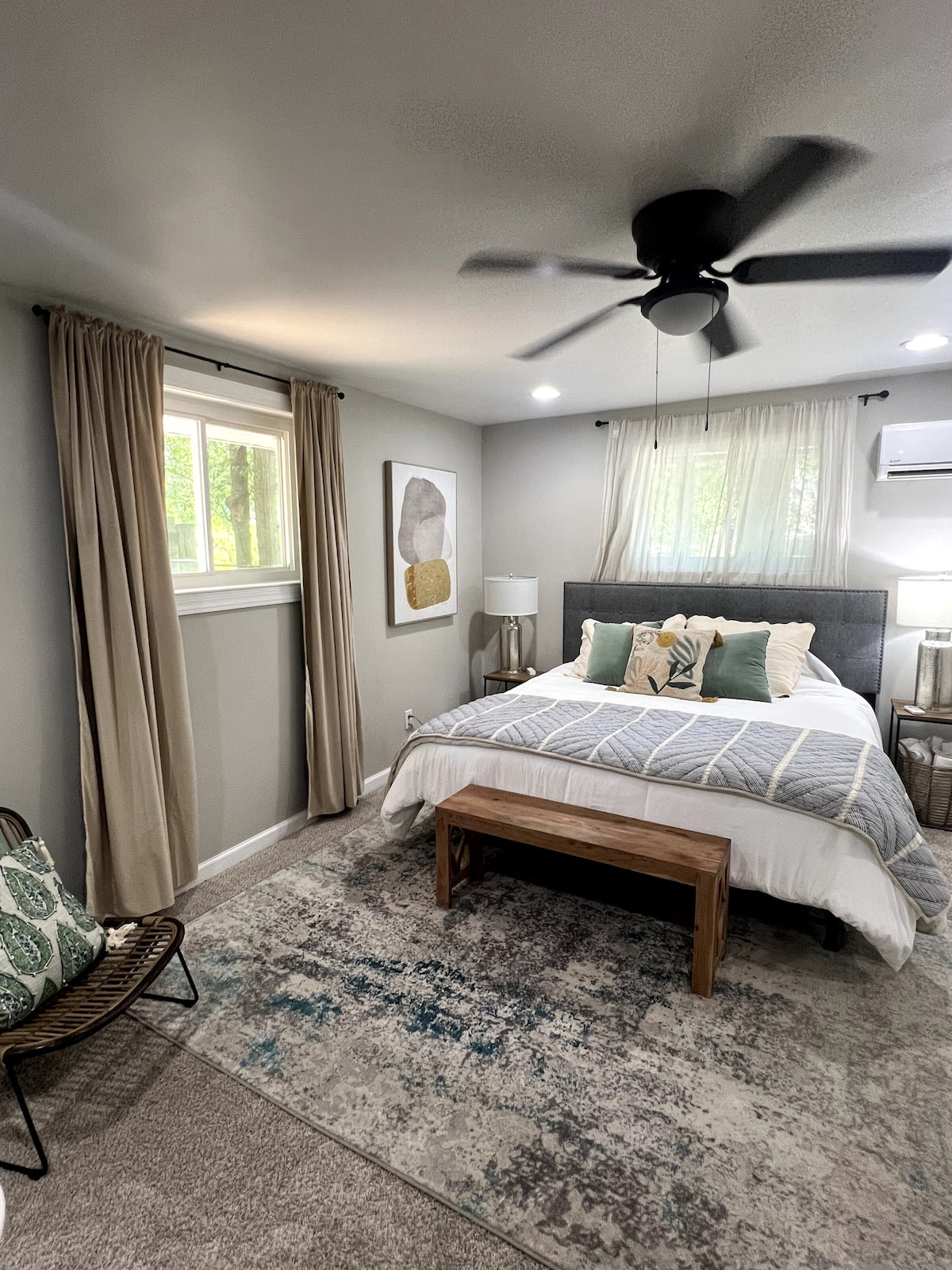 The serene bedroom features a spacious bed adorned with soft linens and decorative pillows. Natural light filters through the partially draped windows, while a stylish area rug adds comfort underfoot. A bench and a cozy chair provide additional seating within the welcoming space.